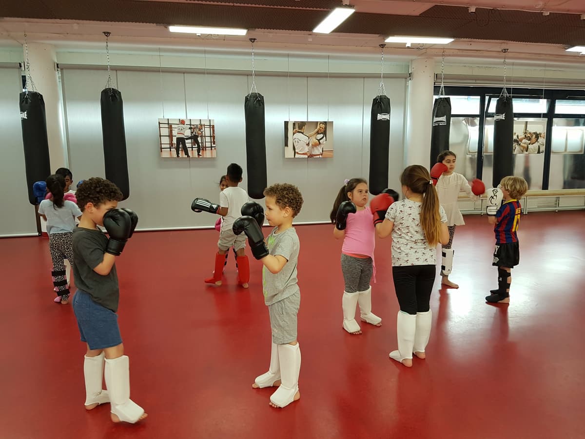 Een groep kinderen met bokshandschoenen traint in een sportzaal met een rode vloer